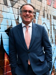 Image of Paul McDonald in a blue suit with pink tie standing in front of a brick wall with graffiti